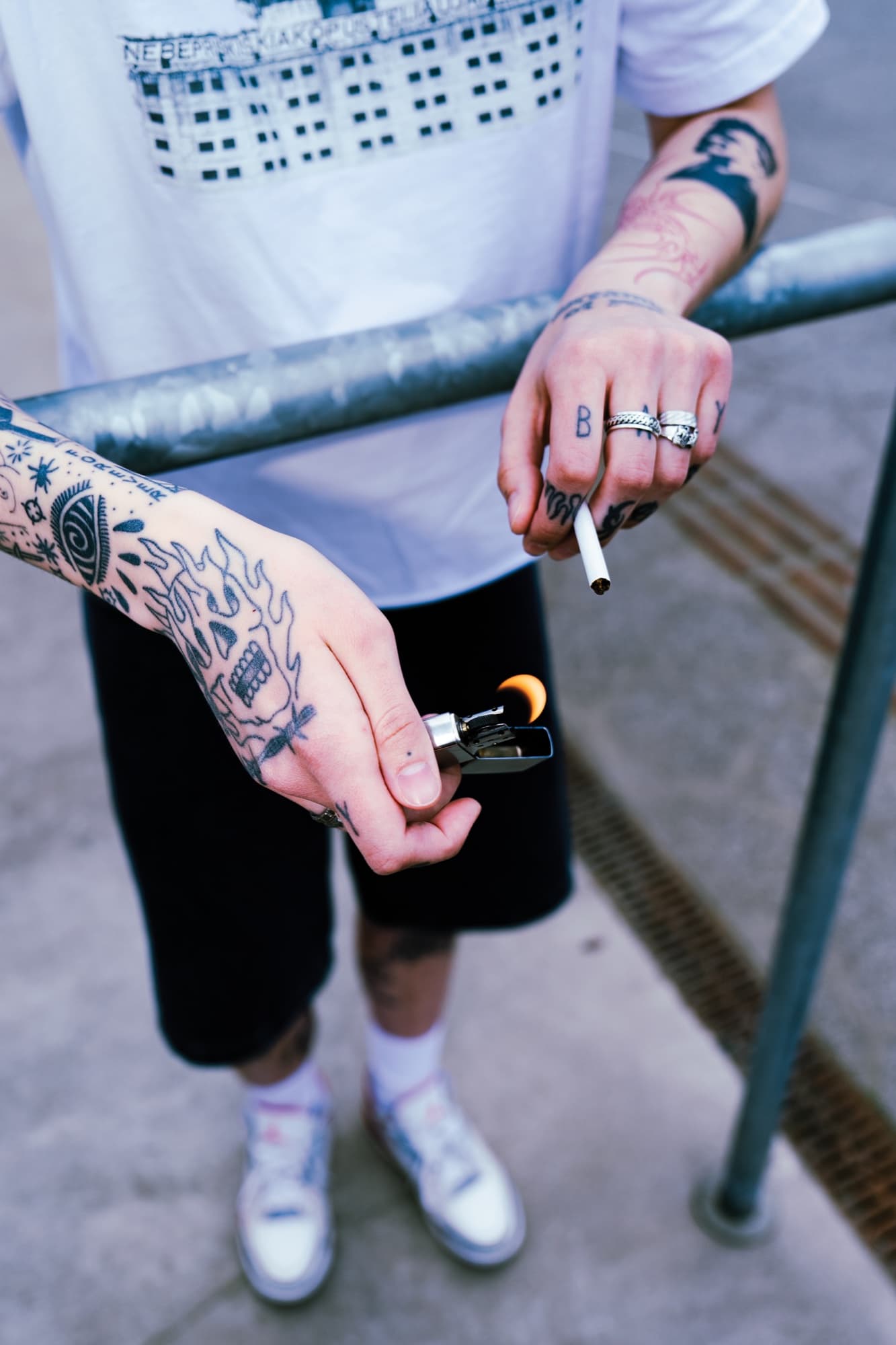 Prie turėklo su Zippo – šortai, balti sportbačiai, blackwork ant dilbio.