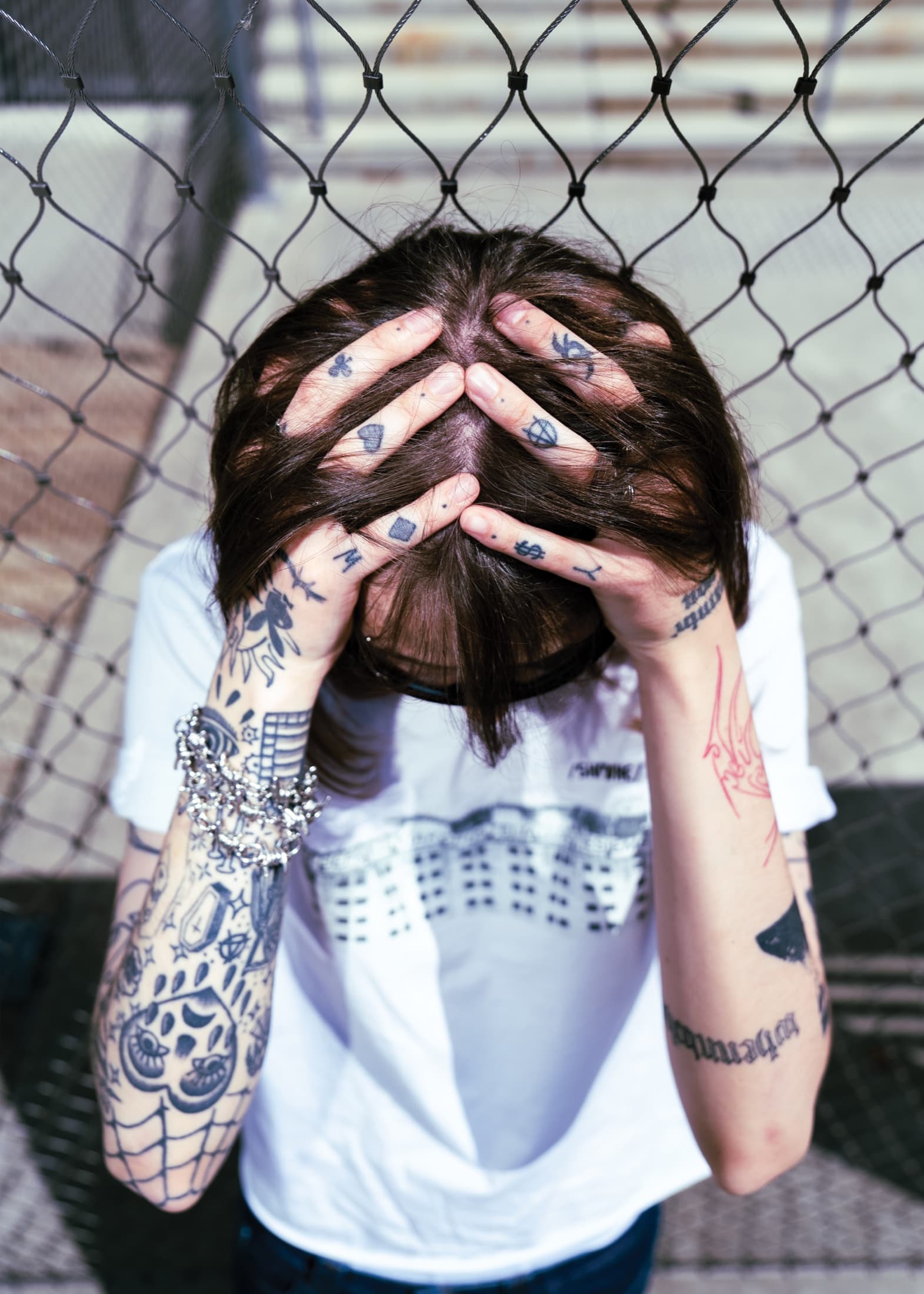 Close portrait against a chain-link fence, tattooed hands over the face.