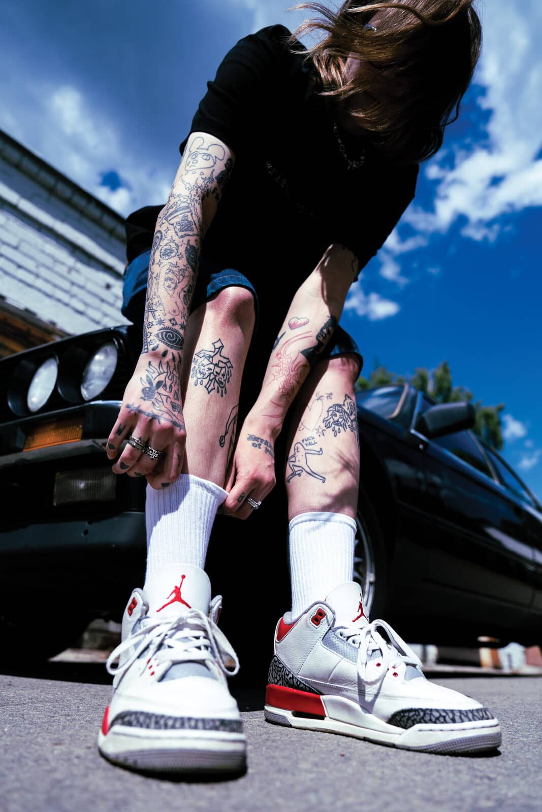 Low angle by the black E30 – tying Jordan 3s, tattooed shins, deep blue summer sky drifting.