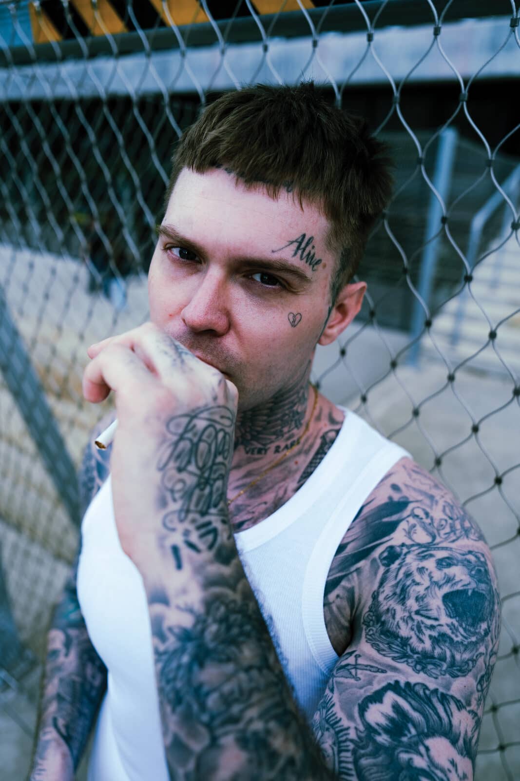 Portrait at the chain-link fence – 'Alive' above the brow, cigarette smoke curling, eyes on the lens.