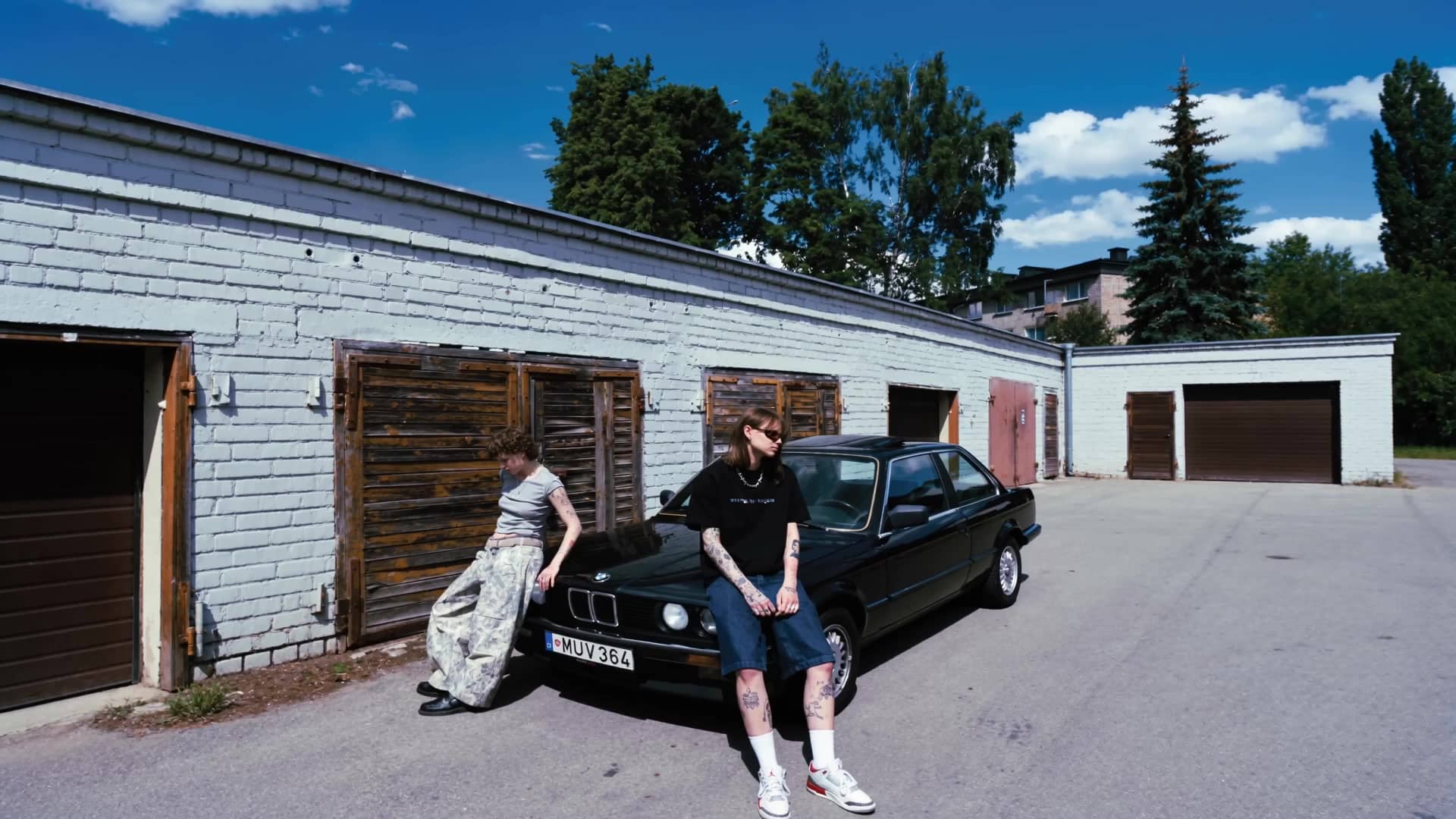 Two figures and a black BMW E30 by the garages – summer air, drifting clouds.